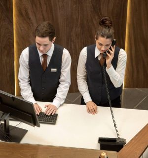 One of two young hotel receptionists standing by counter and looking at computer screen while his colleague answering phone near by